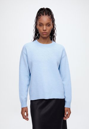 Femme aux cheveux tressés portant un pull bleu clair et une jupe noire, debout devant un fond blanc uni.