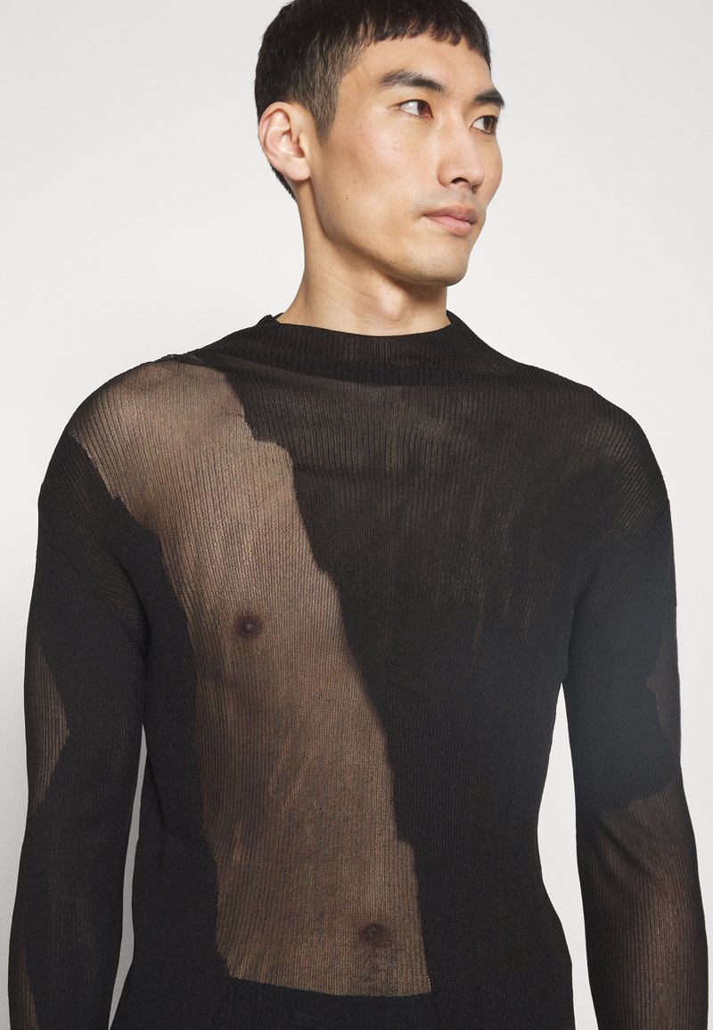 Maglia nera a maglia con sezioni trasparenti, caratterizzata da una texture a costine. Collo a giro e maniche lunghe, che mostrano la pelle sottostante.