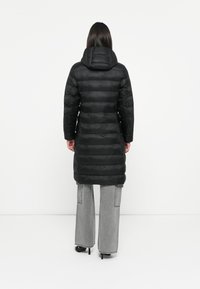 Zwarte pufferjas met capuchon, voorzien van horizontale stiksels, een lange snit en ingezette mouwen, gecombineerd met grijze jeans met wijde pijpen.
