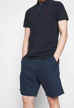 Homme portant un polo à manches courtes bleu marine et un short assorti bleu marine, les mains dans les poches, sur un fond uni.