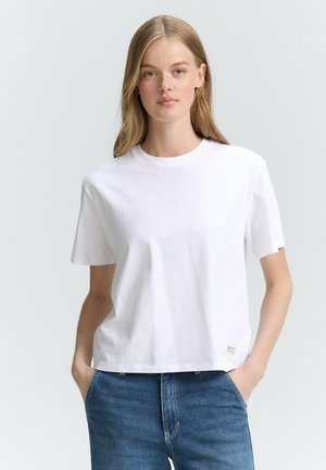 Witte katoenen t-shirt met een ronde halslijn en korte mouwen; heeft een label aan de onderzijde rechts, gecombineerd met blauwe denim jeans.