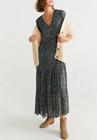 Dunkelblauses Blumen-Maxikleid mit gestuftem Rock und V-Ausschnitt. Cremefarbener gerippter Cardigan mit großen Knöpfen. Beigefarbene Knöchelstiefel runden das Outfit ab.