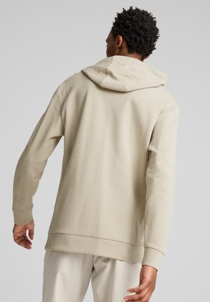 Mężczyzna w jasnobeżowej bluzie z kapturem i pasujących spodniach, stojący tyłem do kamery na jednolitym szarym tle.