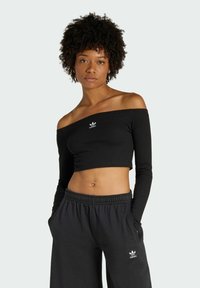 Czarny top typu crop off-the-shoulder z długimi rękawami, z małym białym logo Adidas na piersi. Noszony z czarnymi dresami.