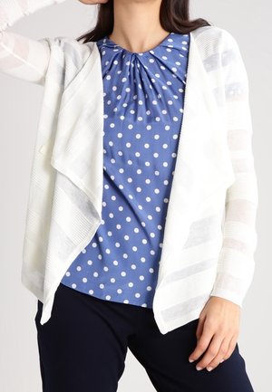 Cardigan en tricot blanc avec un devant ouvert et un motif rayé sur un chemisier bleu à pois, présentant un col froncé et des manches courtes.