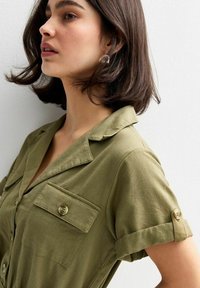 Camisa verde con cuello y mangas cortas, dos bolsillos en el pecho, mangas enrolladas y detalles de botones. Textura suave y tela ligera.