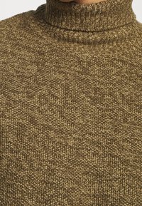 Gros plan sur un pull à col roulé en tricot texturé marron porté par une personne, montrant les motifs de points détaillés et la texture du tissu.