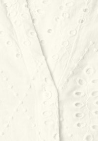 Tissu blanc avec broderie à œillets présentant des motifs floraux et circulaires, plié avec un bouton visible et des détails de couture texturés.