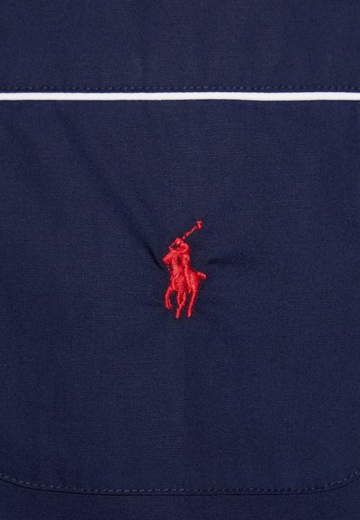 Navy blue fabric features a prominent red embroidered polo player logo with a mallet. Smooth texture with white stitching detail above.