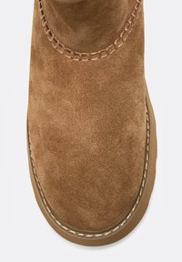 Brązowy zamszowy botek za kostkę z zaokrąglonym noskiem, ozdobnym przeszyciem wzdłuż kołnierza i teksturowaną powierzchnią. Lekka gumowa podeszwa.