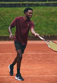 Maroon urheilupaita, mustat shortsit ja mustat kengät valkoisilla yksityiskohdilla. Henkilö, joka pitää tennismailaa savikentällä.