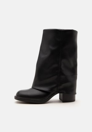 Botte en cuir noir mi-mollet avec un bout arrondi, un petit talon bloc et une texture douce légèrement froissée sur fond blanc.