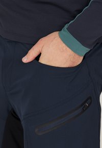 Marineblaue Softshell-Hosen mit einem kontrastierenden türkisfarbenen Saum, ausgestattet mit einer Seitenreißverschlusstasche und einer glatten Textur, geeignet für Outdoor-Aktivitäten.