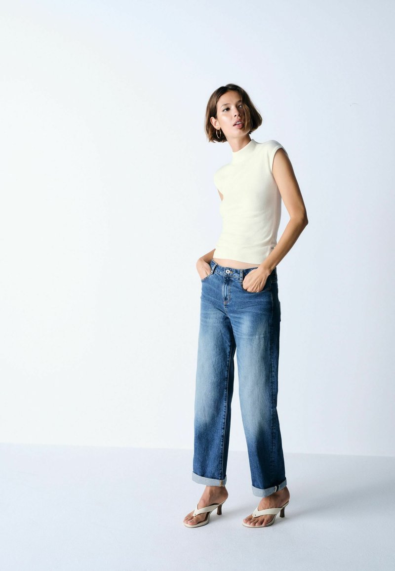 Mujer con camiseta blanca sin mangas, vaqueros azules con puños y sandalias blancas de tacón, de pie con las manos en los bolsillos frente a un fondo claro y liso.