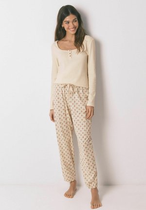 Kvinde står barfodet iført en cremefarvet langærmet top og beige blomstrede løse bukser med snøre, smilende og kigger til siden.