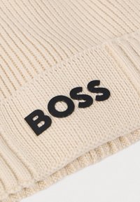 Bonnet tricoté en crème avec le logo "BOSS" noir. Présente une texture côtelée et un ourlet roulé, fabriqué à partir d'un matériau doux et chaud.