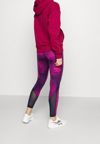 Tighta lila leggings med ett texturerat mönster, svarta mesh-detaljer, kombinerat med en vinröd hoodie och vita träningsskor.