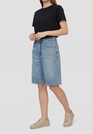 Donna che indossa una maglietta nera a maniche corte, pantaloncini di jeans azzurri fino al ginocchio con bottoni laterali e sneaker beige, in piedi con le mani giunte.