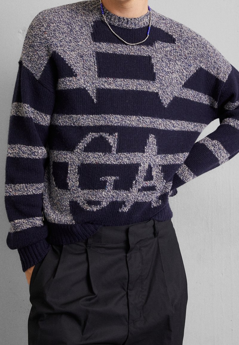 Personne portant un pull en maille bleu marine et gris à motifs avec des rayures horizontales et des formes abstraites, associé à un pantalon plissé noir.