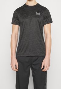 Černé sportovní tričko s vlnkovým vzorem, krátkými rukávy, kulatým výstřihem a bílým logem na levém prsu. V kombinaci s černými kalhotami.