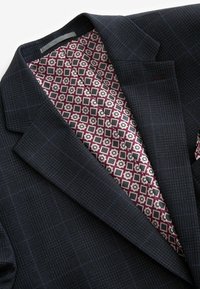 Blazer bleu foncé avec un tissu texturé au motif à carreaux subtil ; doublure géométrique bordeaux et grise.