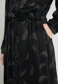 Svart klänning i ett mjukt, texturerat tyg med midjeband. Har ett subtilt guldmönster och knappar framtill samt sidofickor.