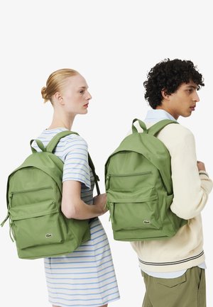 Dos personas con mochilas verde claro con bolsillos frontales, de pie lado a lado, mirando en direcciones opuestas sobre un fondo blanco.