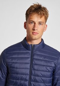 Marinblå puffjacka med kviltat mönster, hög krage och framzip. Släta, lätta material med horisontella sömmar överallt.