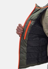 Giacca isolante verde con rifiniture nere, caratterizzata da una zip arancione e dettagli arancioni. Tasca interna con chiusura a zip e texture trapuntata.