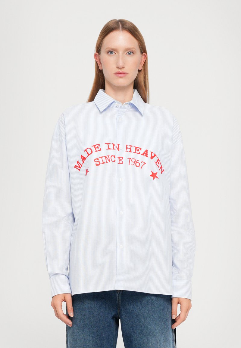 Camisa azul e branca às riscas com colarinho abotinado, apresentando texto bordado em vermelho "FEITO NO CÉU DESDE 1967" e estrelas.