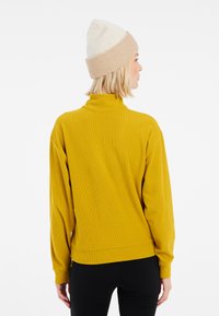 Langarm-Turtleneck-Pullover in Senfgelb, strukturierter Stoff, lockere Passform und gerippte Bündchen; kombiniert mit schwarzen Leggings und einer beige gestrickten Mütze.