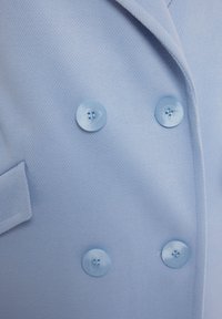 Jasnoniebieska dwurzędowa marynarka wykonana z fakturowanej tkaniny, z dużymi dopasowanymi guzikami i jedną kieszenią.