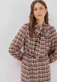 Braunes Karokleid mit einem Bindekragen, langen Ärmeln und einer taillierten Taille. Der Stoff zeigt eine Mischung aus dunklen und hellen braunen Quadraten und Rechtecken.