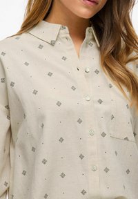 Camisa beige de botones en una tela ligera, con un sutil patrón de diamantes y acentos negros. Incluye un bolsillo y un cuello estructurado.