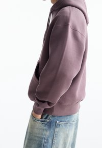 Felpa di cotone viola con tasca a canguro e cappuccio con coulisse, indossata sopra jeans di denim azzurro chiaro. Presenta polsini e orlo a coste.