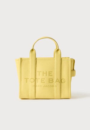 Dzeltena ādas somiņa ar tekstūru, divi augšējie rokturi, iepresēts teksts "THE TOTE BAG" un "MARC JACOBS", strukturēta forma.
