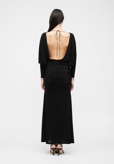 Γυναίκα με μακριά σκούρα μαλλιά φοράει μαύρο φόρεμα μήκους μέχρι το πάτωμα με χαμηλή ανοιχτή πλάτη δεμένη στο λαιμό, στέκεται με την πλάτη στον θεατή.