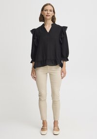 Blusa nera con maniche a sbuffo, scollatura a V e tessuto testurizzato; abbinata a pantaloni beige attillati e scarpe di colore chiaro.