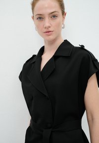 Robe noire à ceinture avec un devant double boutonnage, un large col et des manches courtes. Tissu lisse avec des détails de boutons sur les épaules.