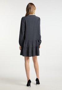 Schwarzes, langärmliges Kleid mit einem gestuften Rock und einer glatten Textur. Das Design verfügt über einen subtilen Puff an der Schulter und einen Rückenverschluss.