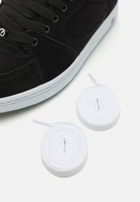 Must musta sneakersid, millel on tekstuuriga ülemine osa, valge kummitald ja lamedad mustad paelad. Kaks valget ümmargust paela on asetatud lähedusse.