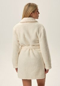 Kremowy polarowy szlafrok z kołnierzykiem, długimi rękawami i paskiem w talii; miękka faktura o lekko puszystym wyglądzie.