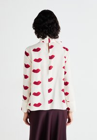 Blouse à manches longues en tissu crème avec un motif de lèvres rouges répétées. Détail en forme de clé à l'arrière et fermeture par bouton au cou.
