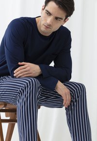 Camisa de manga larga azul marino con un bolsillo en el pecho y pantalones de pijama a rayas blancas. Sentado en una silla de madera contra un fondo claro.