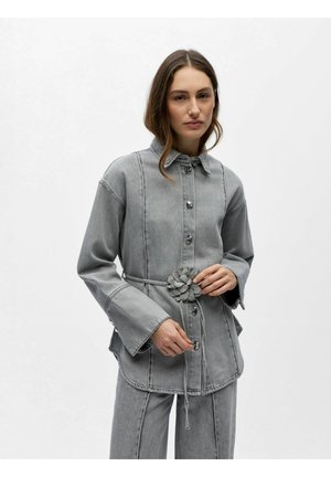 Ženska v sivi gumbeni jopiči iz trapera z razširjenimi rokavi in ujemajočimi hlačami, ki v pasu drži tkan pas z rožami.