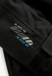 Tessuto tecnico nero con testo stampato riflettente e blu. Presenta una chiusura in velcro e linee che indicano misure e coordinate.