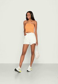 Top halter laranja com um design ajustado, combinado com calções de ganga brancos de cintura alta e ténis brancos com realces neon, segurando uma bolsa de vime.