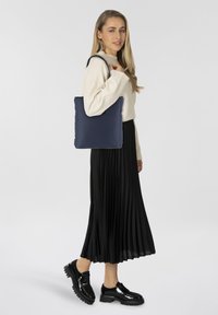 Sac de tote matelassé bleu marine avec des poignées plates, associé à un pull crème et une jupe plissée noire. Des chaussures vernies noires à lacets complètent le look.