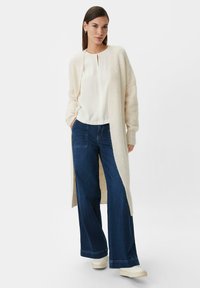 Long, cream-colored cardigan made of soft material, paired with a white blouse and blue wide-leg jeans, and white sneakers.