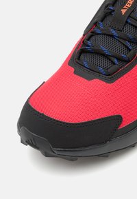 Roter und schwarzer Sportschuh mit Mesh-Obermaterial, strukturiertem Oberfläche, gepolsterter Zunge und blauen Akzenten an den Schnürsenkeln. Verfügt über eine robuste schwarze Sohle.
